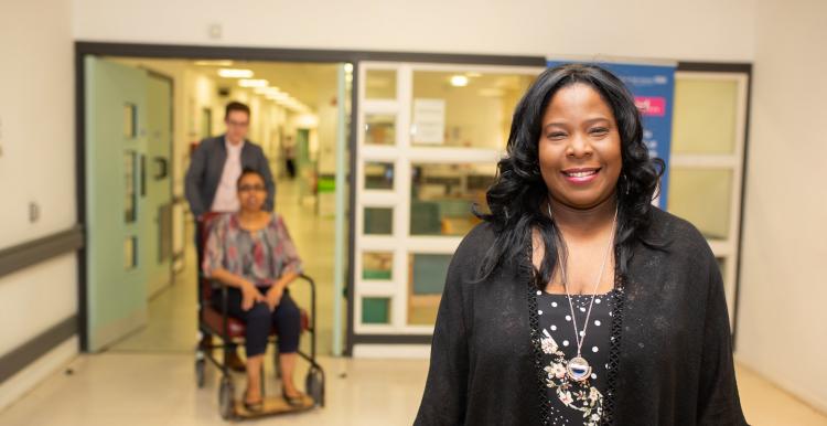 Woman in hospital smiling at the camera with a man pushing a woman in a wheelchair in the background